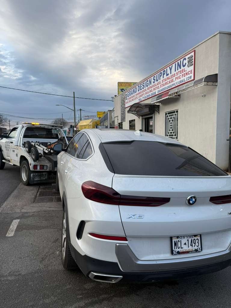 White BMW X6 being towed past Grand Design Supply on a Queens commercial street — JG Towing.