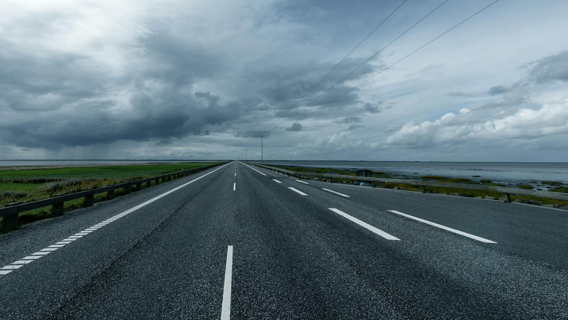 A straight road stretches toward the horizon under dramatic cloudy skies along a coastal landscape.